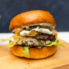 A smash burger with tarragon and black truffle, served in a toasted brioche bun with visible layers of meat, cheese, and vegetables.